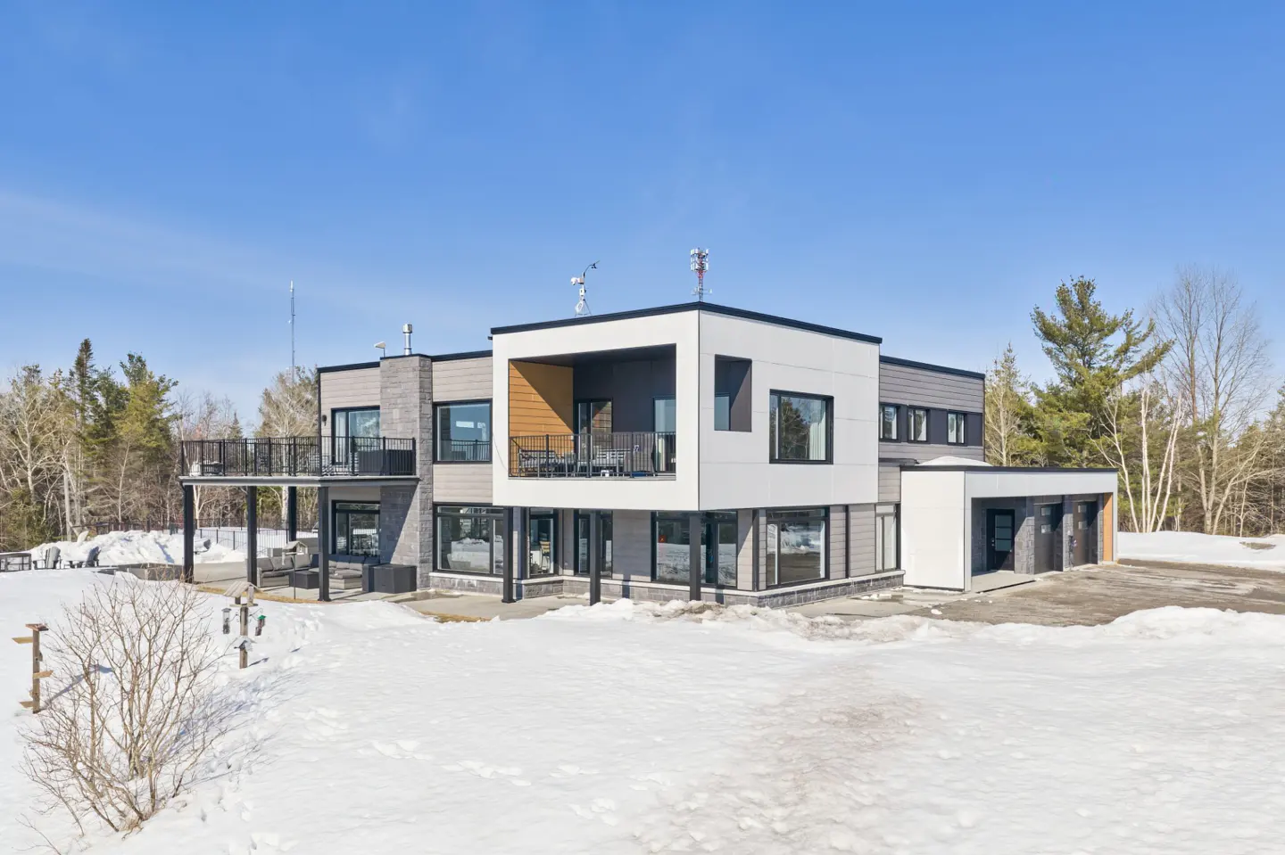 Modern two-story home with a white and gray exterior, black trim, and a balcony, surrounded by snow and trees under a blue sky.