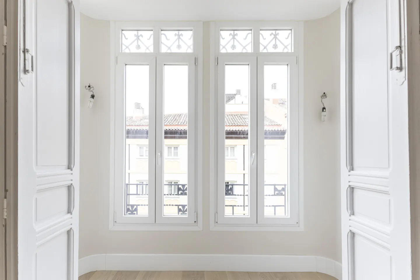 Bright room with open white doors and two white windows. Outside view of buildings. Light wood floor.