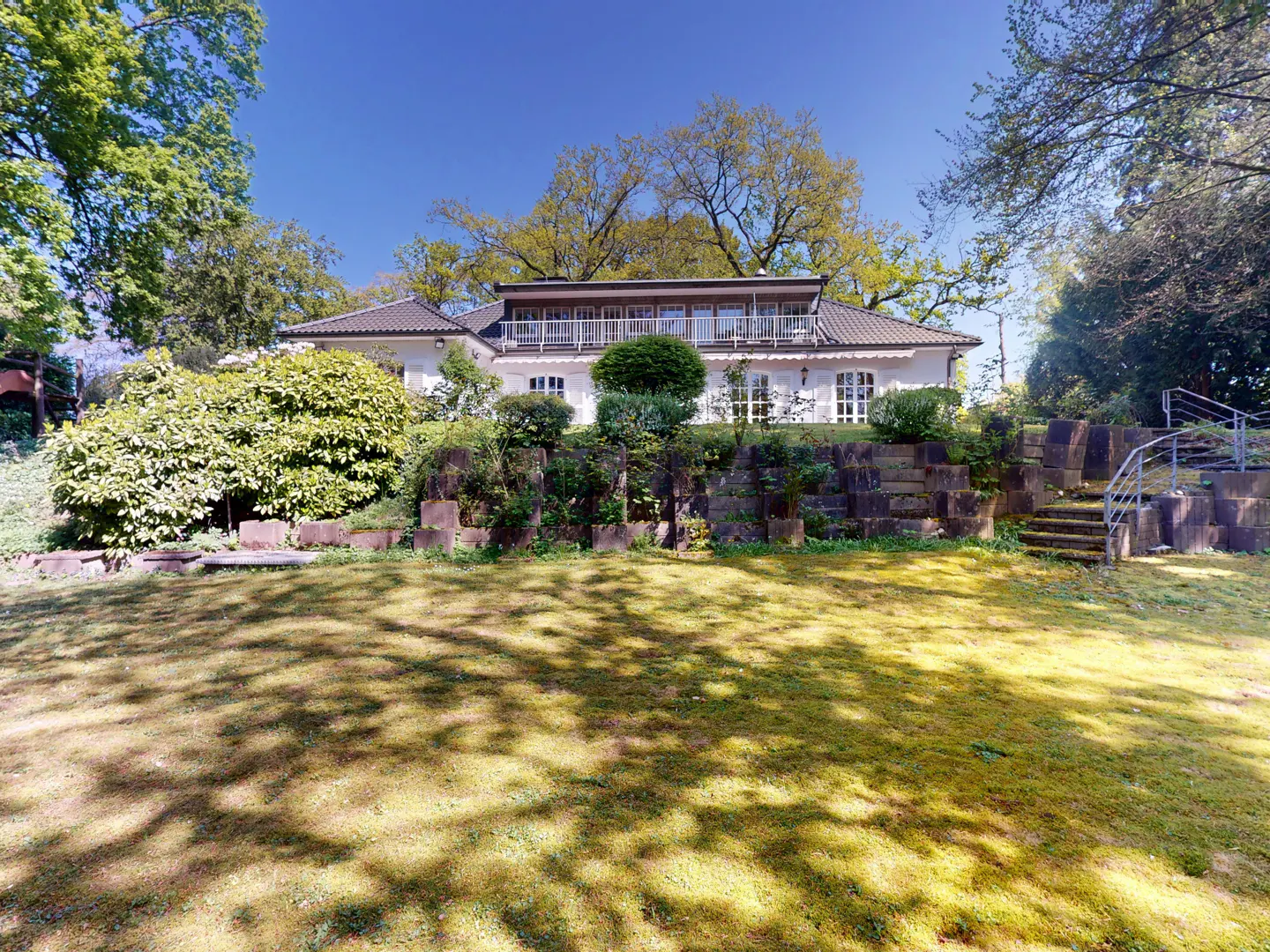 Exterior view of a white house with a balcony, set on a hill with a stone retaining wall and green lawn.