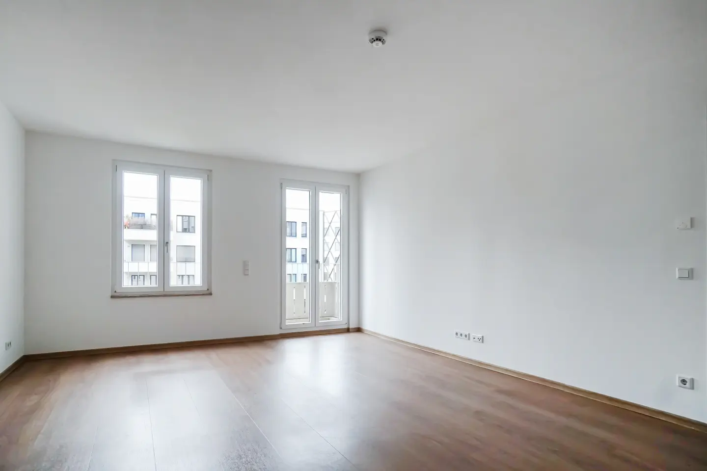Bright, empty room with wood floors, white walls, and two white-framed windows showing a building exterior.
