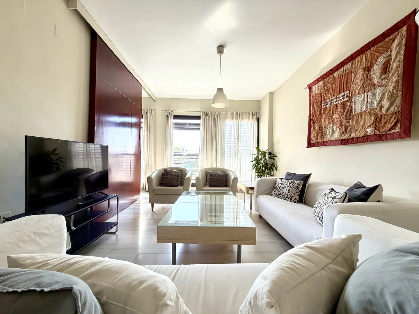 Bright living room with white sofas, armchairs, and a glass coffee table. A large tapestry hangs on the wall. A TV sits near a sliding wood door.