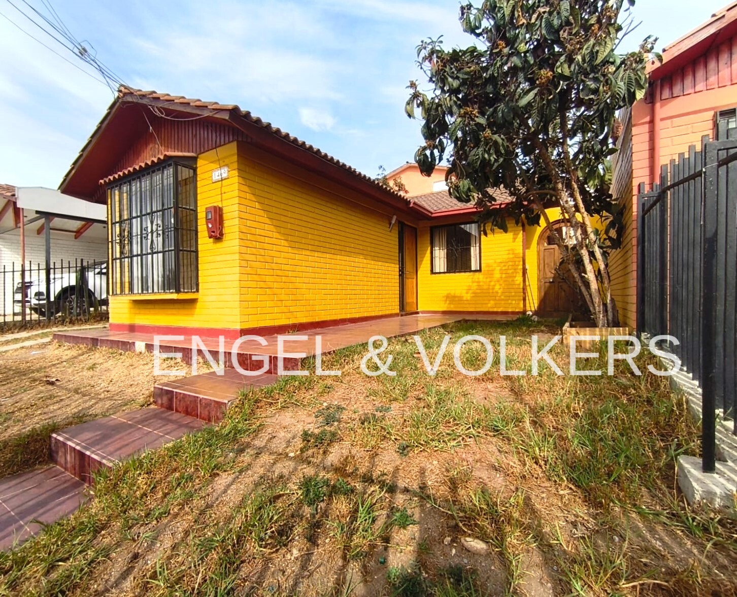 Exterior view of a bright yellow brick house with a tiled roof, a bay window, and a small front yard with a tree.