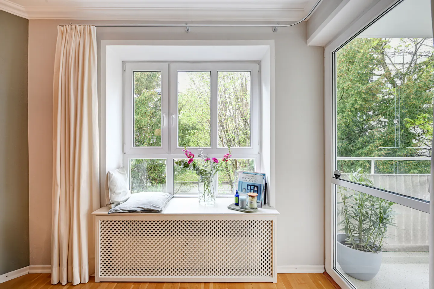 Bright room with a white window seat, pillows, flowers, and books. Cream curtains hang to the left of the window. A glass door leads to a balcony with green trees.
