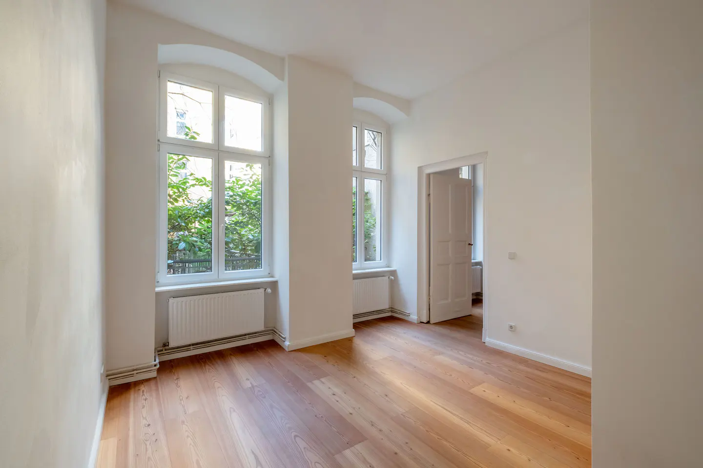 Bright, empty room with hardwood floors, white walls, and two windows with green foliage visible outside. A doorway leads to another room.