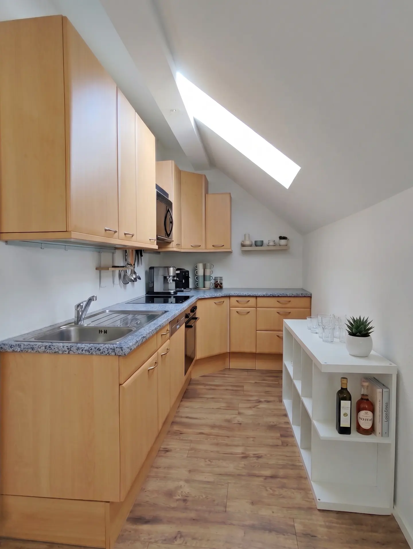 Bright kitchen with wood cabinets, granite counters, and wood floors. A skylight illuminates the room. A white shelving unit holds glasses and decor.