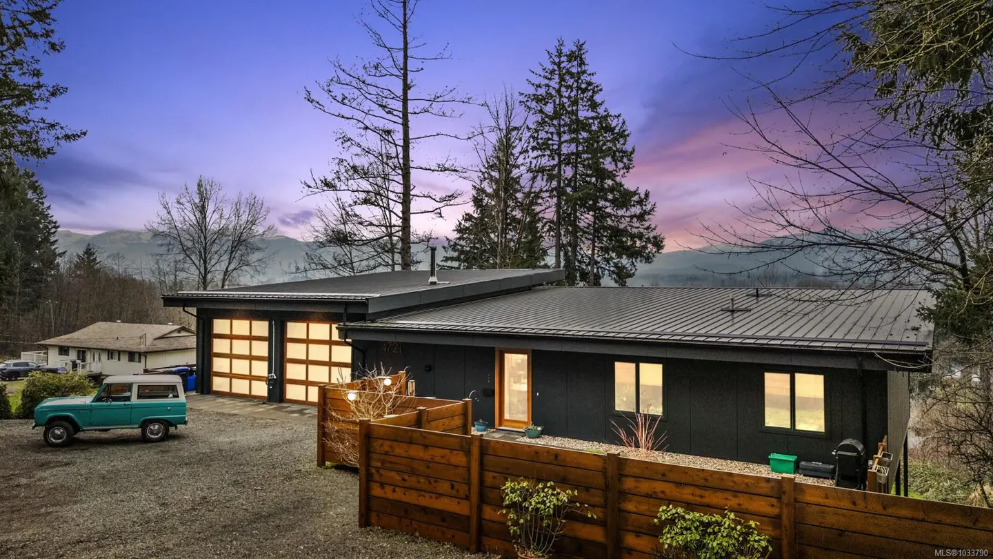 Modern dark gray house with a black roof, wood fence, and a vintage blue Ford Bronco parked in the gravel driveway.