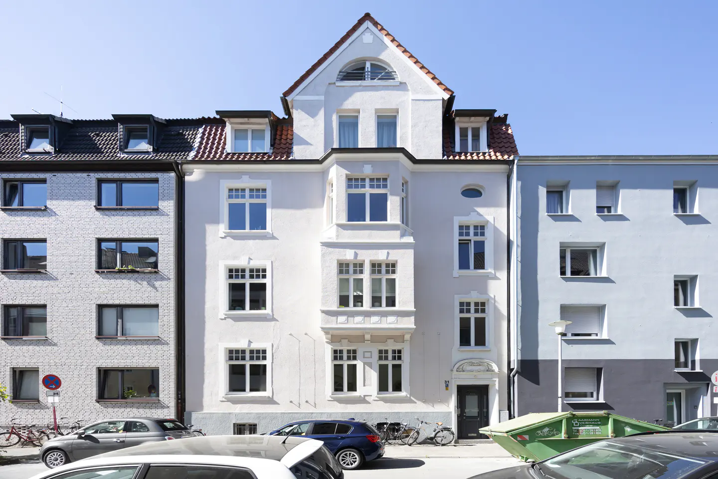 A three-story white building with a red-tiled roof stands between two other buildings on a city street. Cars are parked in front.
