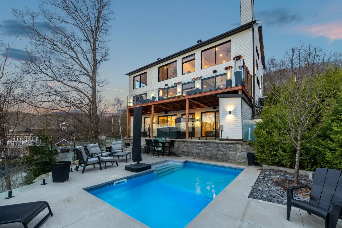 Modern white house with a pool, patio furniture, and a wooden deck overlooking the pool. Mountains are visible in the background.