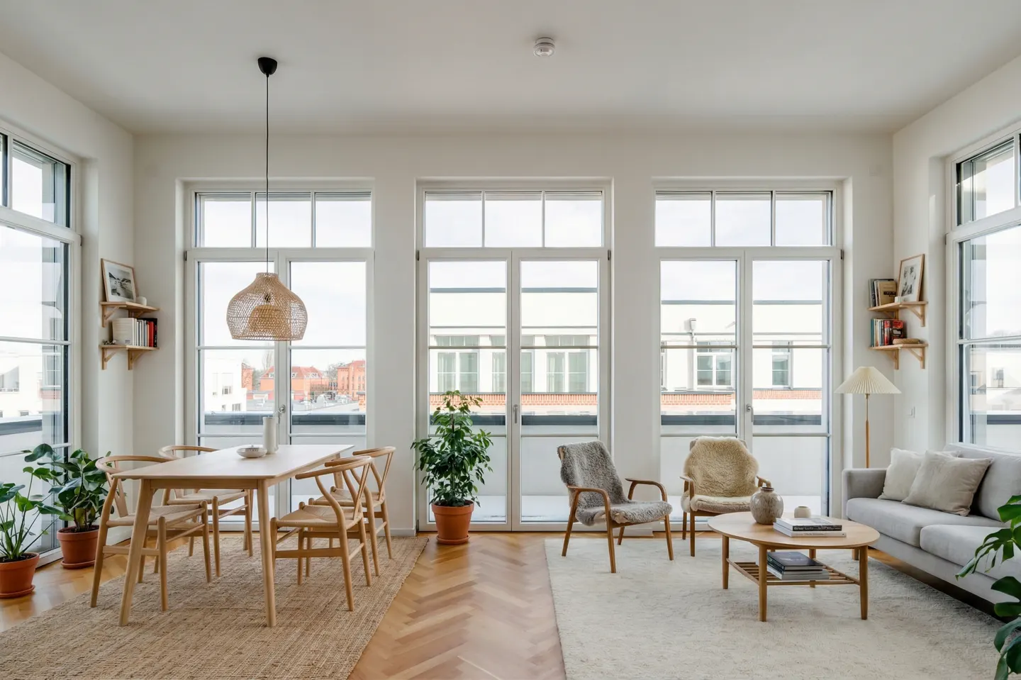 Bright, airy living and dining area with large windows, wood floors, and neutral decor. Table, chairs, sofa, and plants furnish the space.