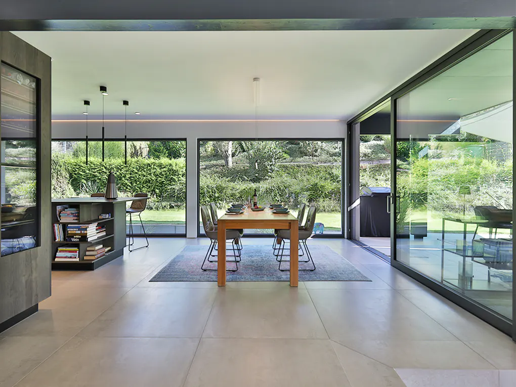 Modern dining room with a wood table set for a meal, surrounded by large windows overlooking a green garden.