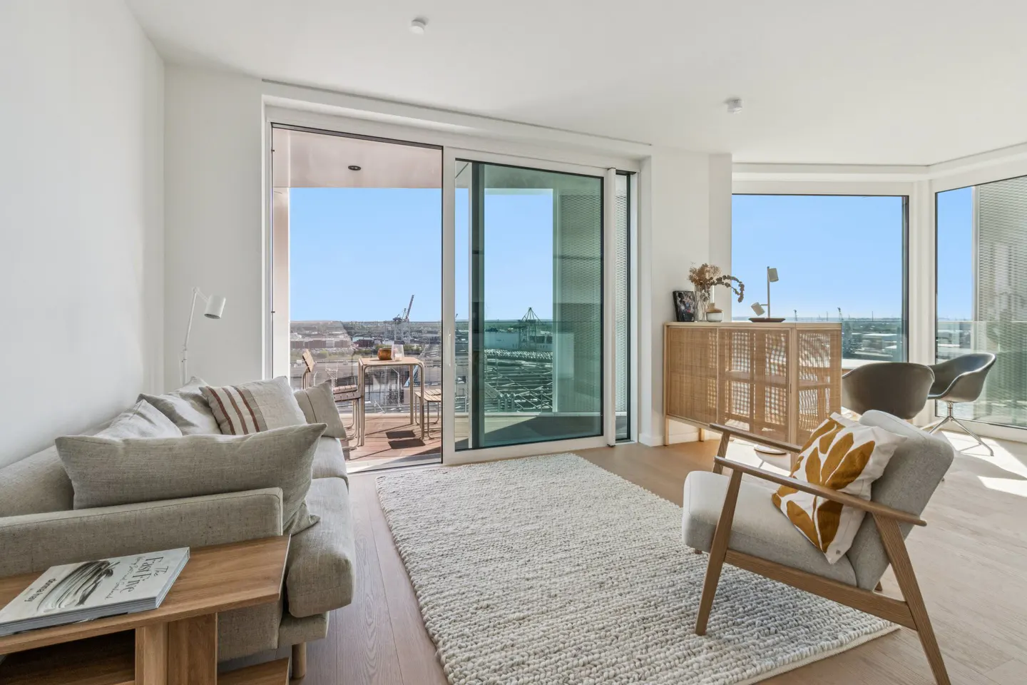 Bright living room with a beige sofa, wooden chair, and a balcony overlooking a cityscape with a harbor.