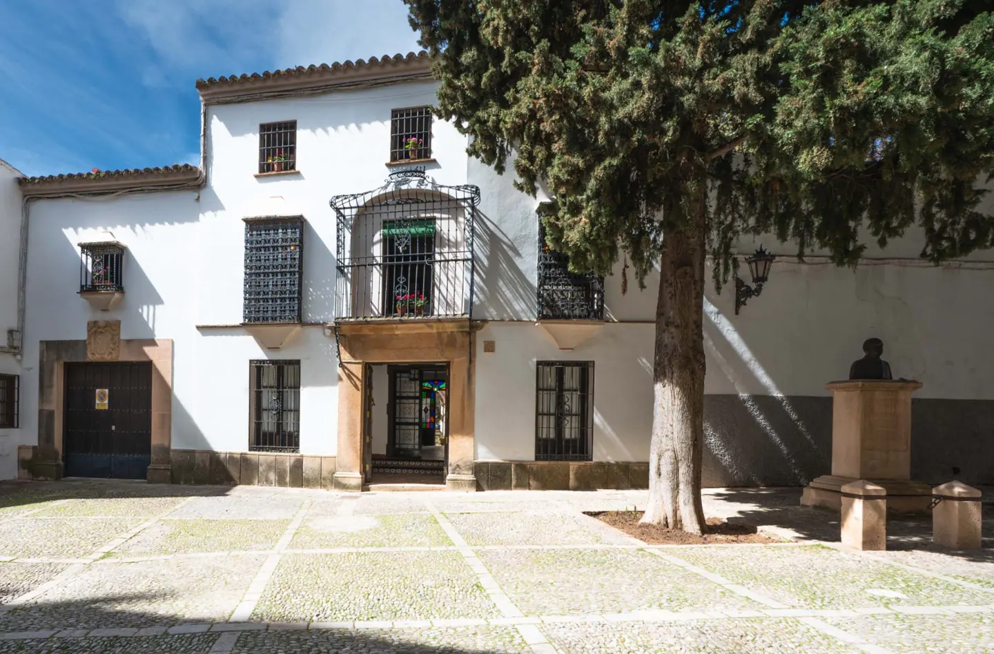 Exterior of a white building with a balcony, dark windows, and a large tree in front. Cobblestone courtyard.