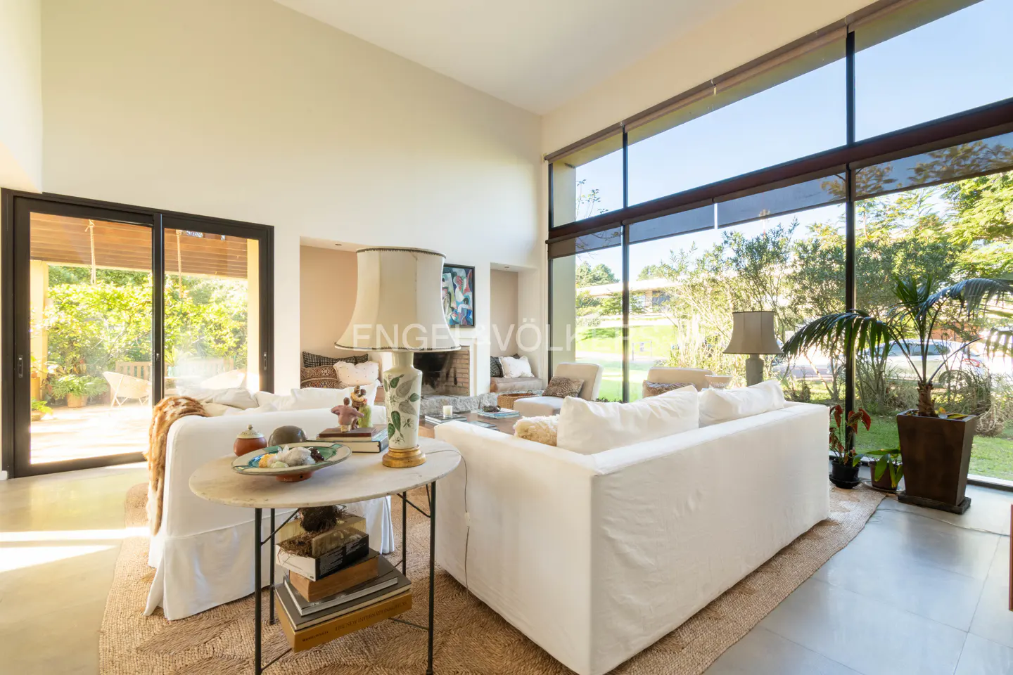 Bright living room with white sofas, a stone table with books, and large windows overlooking a green yard.