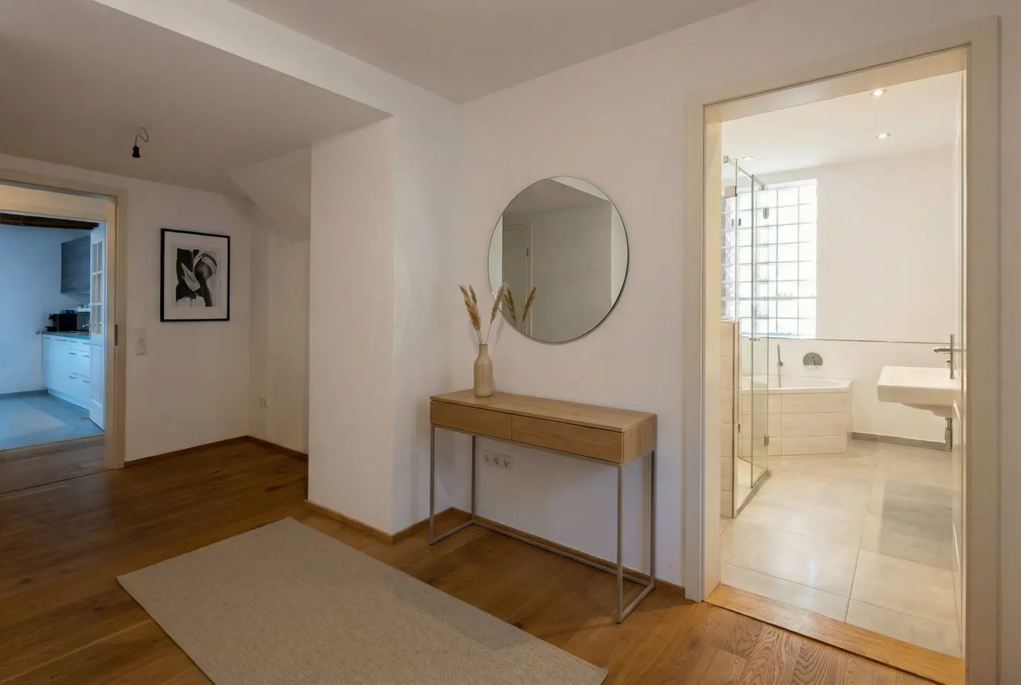 Bright hallway with wood floors, white walls, and doorways to a kitchen and bathroom. A round mirror hangs above a light wood console table.