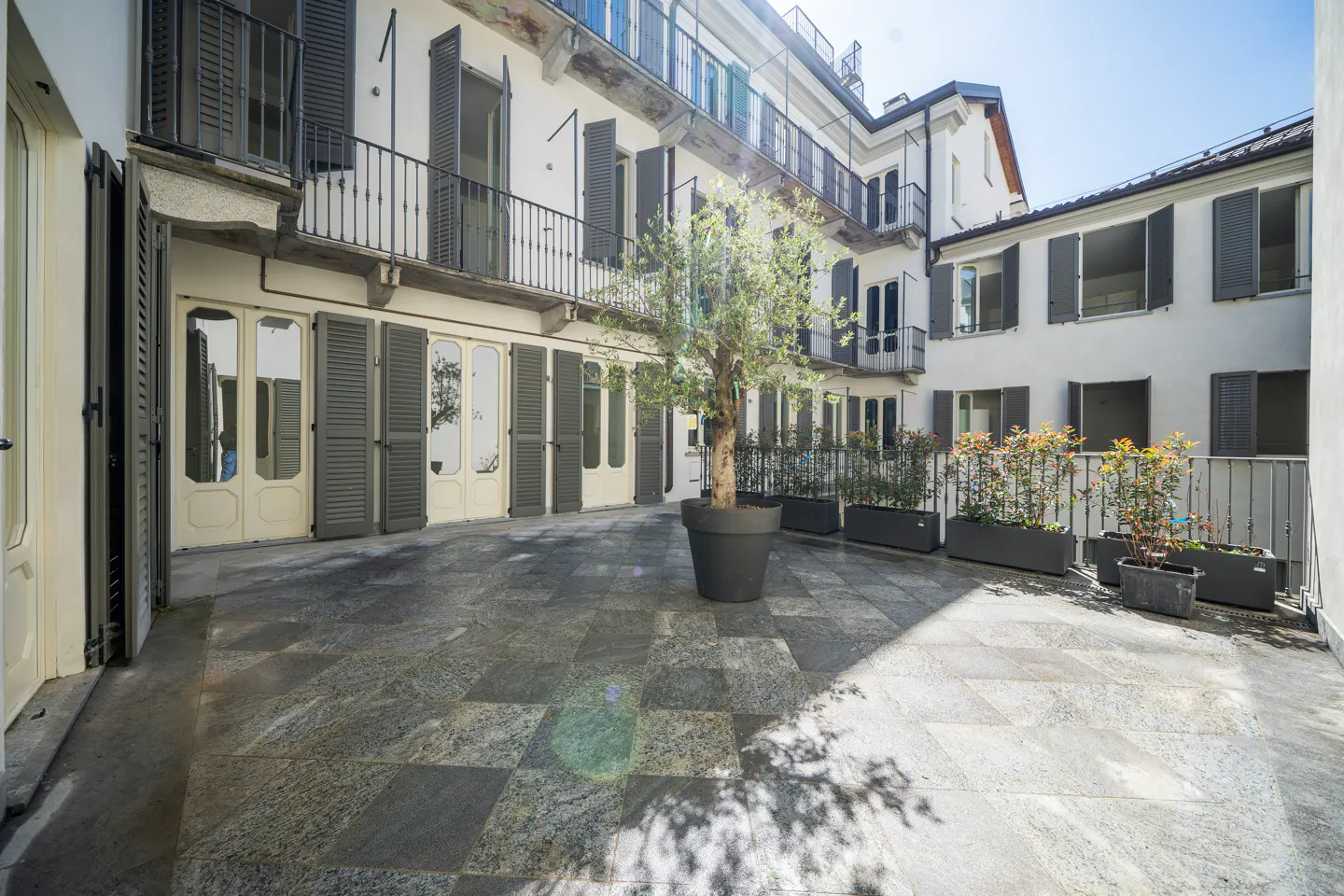 Courtyard view with gray stone tiles, white buildings, and dark gray shutters. A potted tree stands in the center. Balconies and planters add detail.