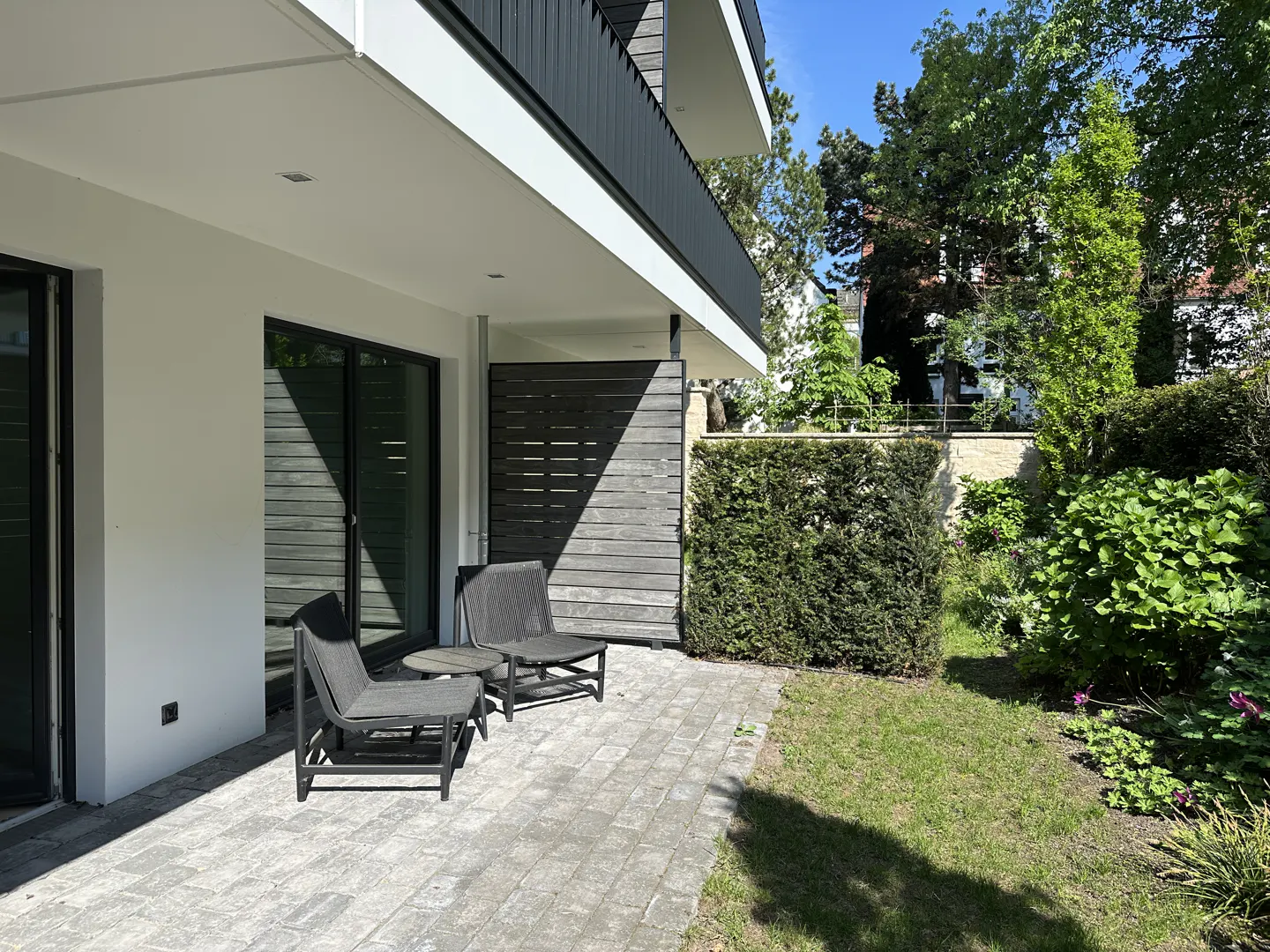 Outdoor patio with two gray chairs and a small table on a stone patio, next to a white building and green lawn.