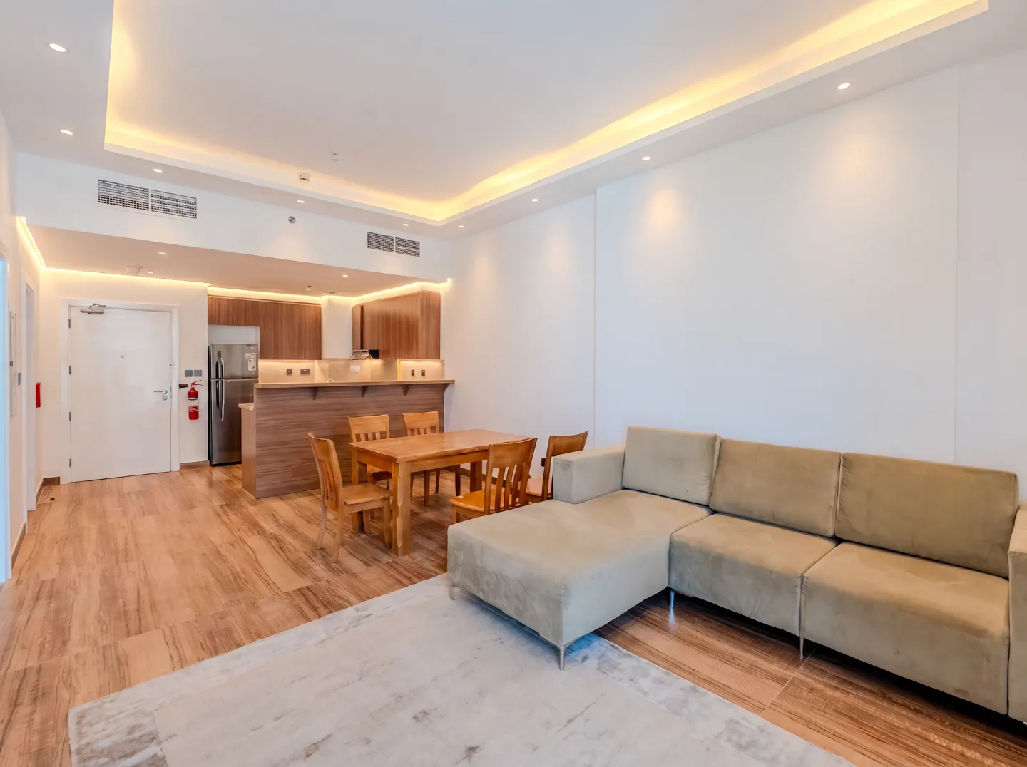 Bright apartment interior with a beige sectional sofa, wooden dining table, and kitchen with wood cabinets.