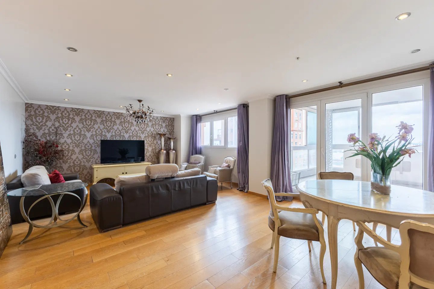 Bright living room with wood floors, brown leather sofas, and a dining table with flowers near large windows with purple curtains.