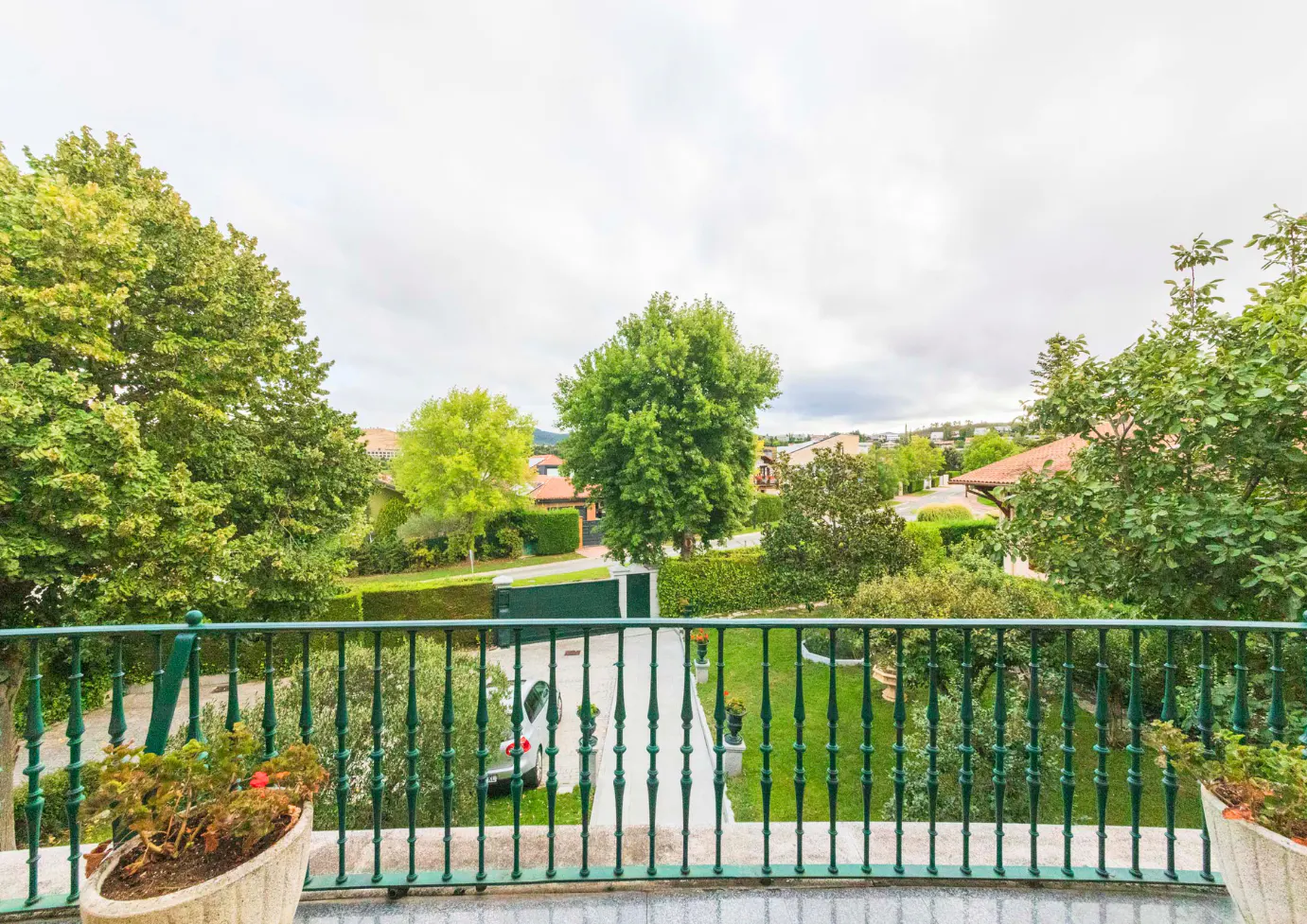 View from a balcony with green railings overlooking a driveway, lawn, trees, and houses under a cloudy sky.