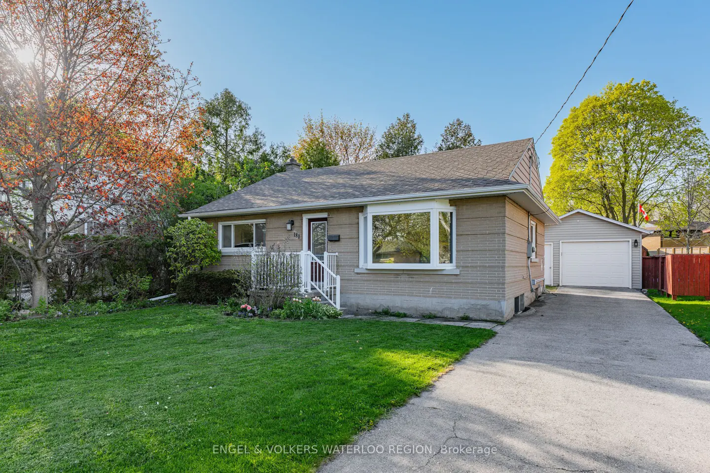 A tan brick bungalow with a gray roof, white trim, and a green lawn on a sunny day. A detached garage is visible.