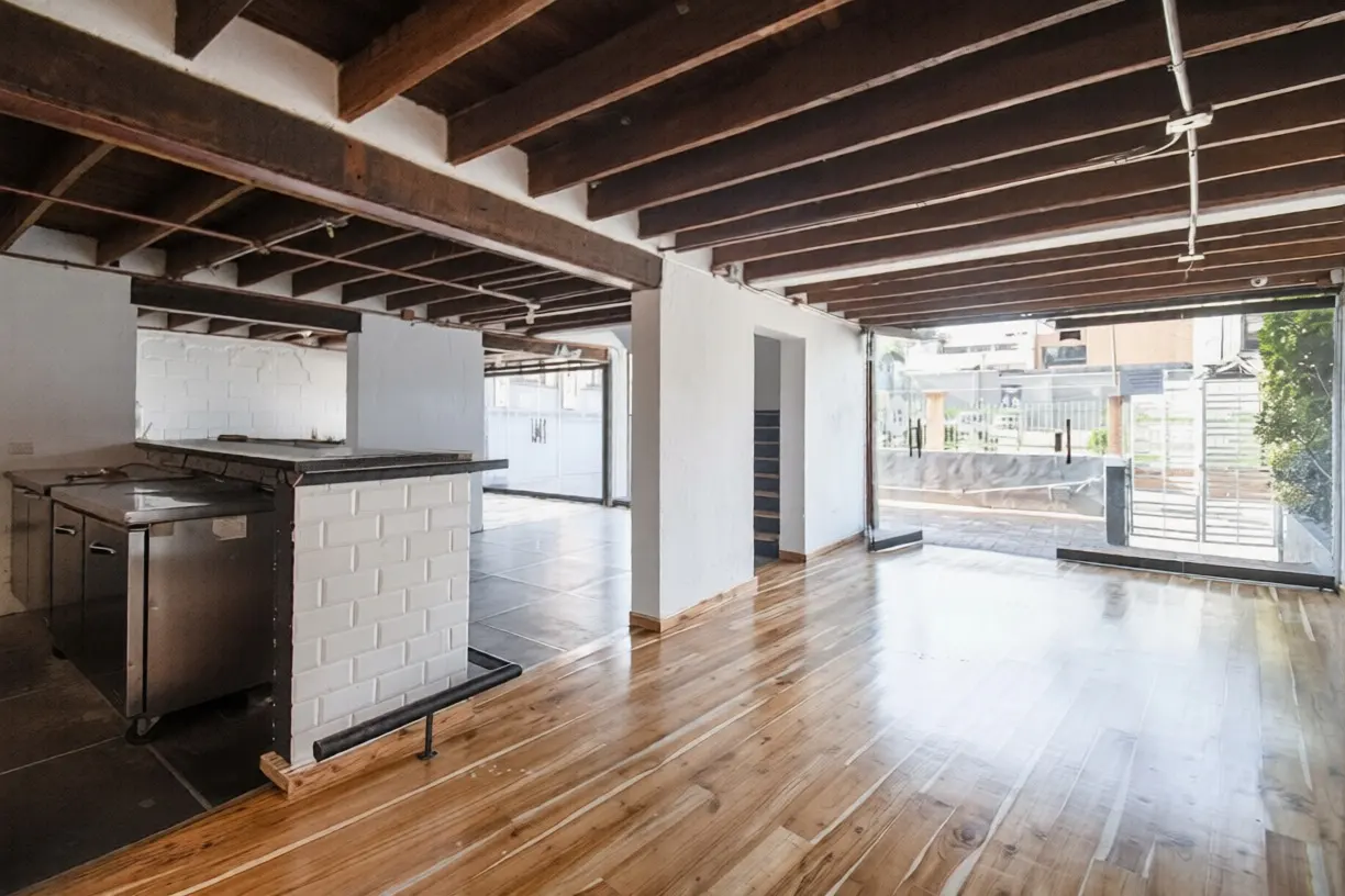 An open-concept room with wood floors, exposed beams, and a white brick bar. Stainless steel cabinets are visible. Large windows offer outdoor views.