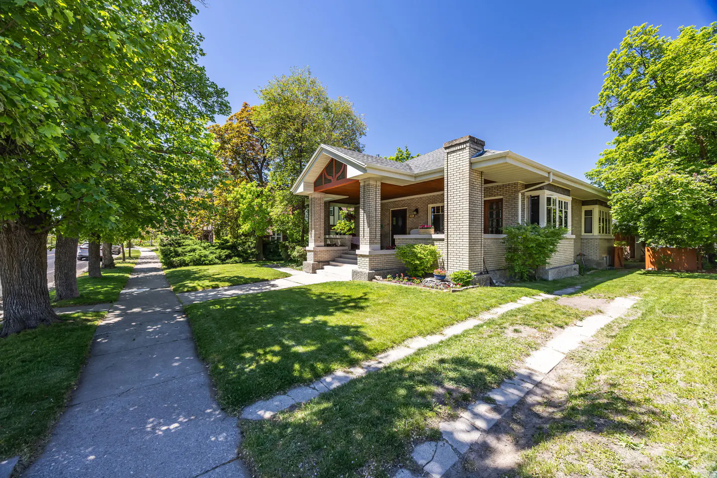 A tan brick house with a porch sits on a green lawn under a blue sky. Trees line the sidewalk.
