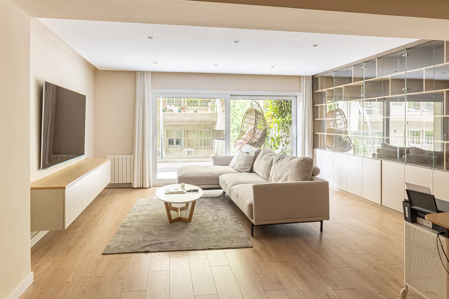Living room with a beige sectional sofa, round coffee table, and a large window with a hanging chair. Wood floors and a mirrored shelving unit.