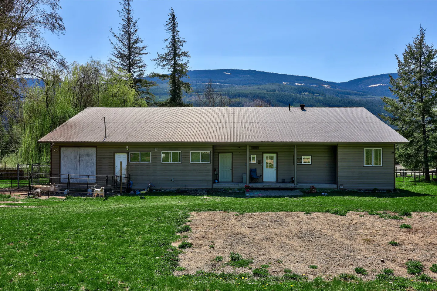 A single-story, gray house with a metal roof sits on a green lawn against a mountain backdrop.