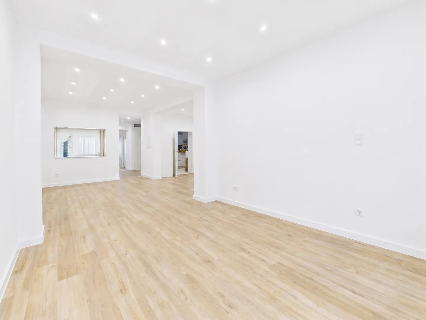 Bright, empty room with light wood floors and white walls. Recessed lighting illuminates the space, which opens to other rooms.