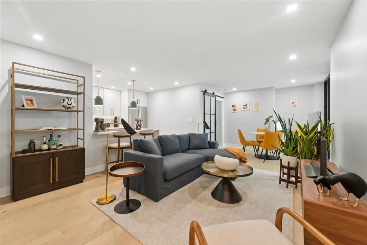 Open-concept living space with a gray sectional sofa, round coffee table, and a dining area with yellow chairs. A bookshelf and kitchen are visible.