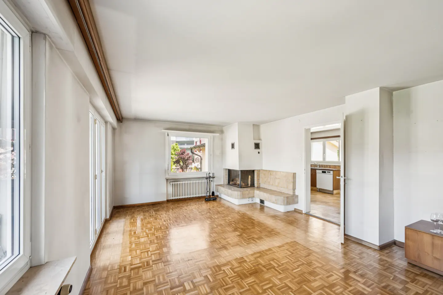 Bright, empty living room with parquet floors, white walls, and a modern fireplace. A window and doorway offer views to the outside and kitchen.