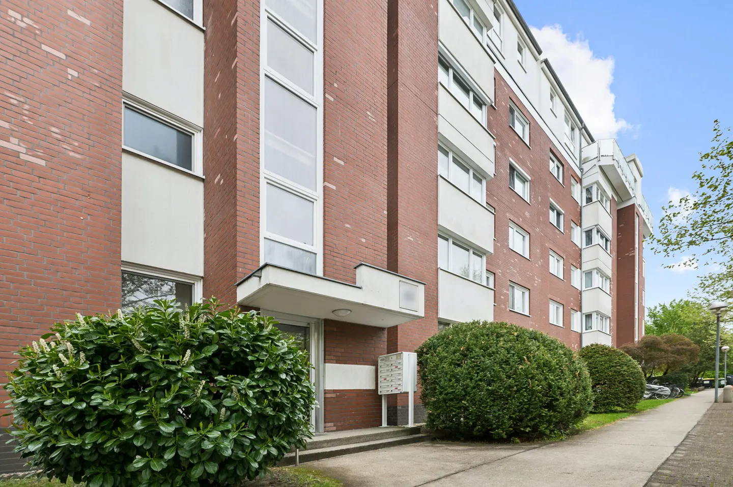 Exterior view of a multi-story red brick apartment building with white accents, a concrete entrance, and green bushes.