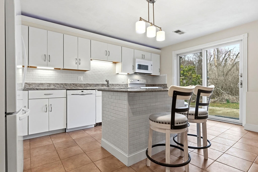Bright kitchen with white cabinets, subway tile backsplash, and a granite countertop island with two bar stools. Sliding glass doors lead to a backyard.