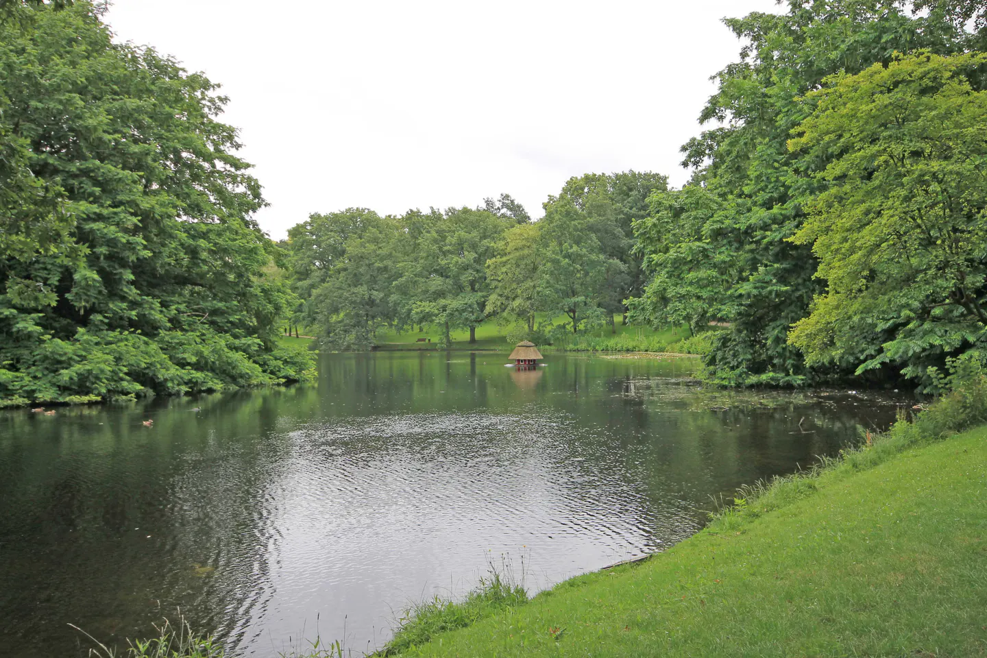 Scenic view of a calm lake surrounded by lush green trees and grass. A small gazebo sits in the middle of the water.