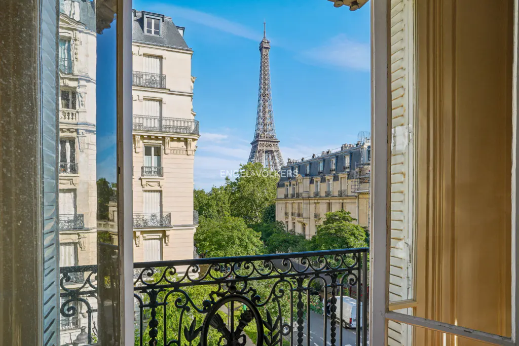 View from a Paris apartment window, showing the Eiffel Tower in the distance. Ornate black iron balcony railing. Blue sky.