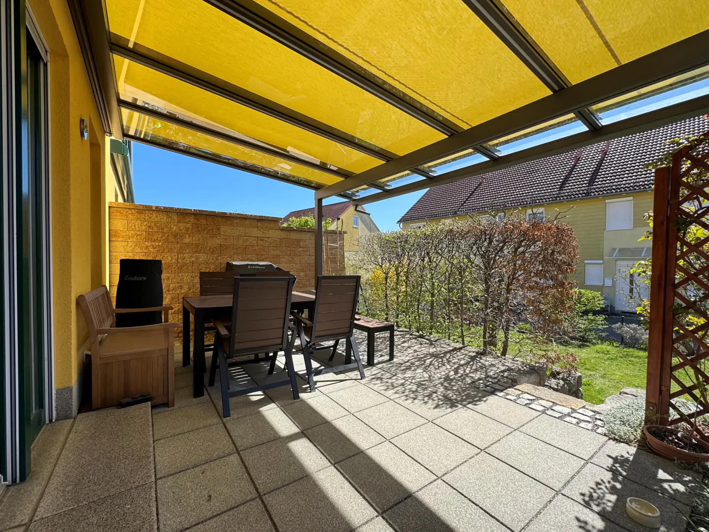 Covered patio with a yellow awning, stone tile floor, and outdoor dining set. A brick wall and green hedge border the space.