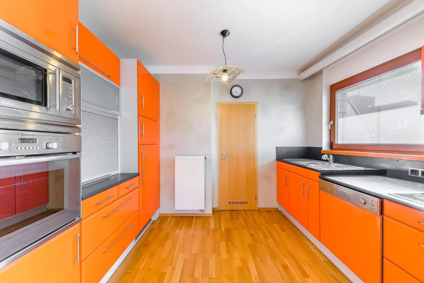 Bright kitchen with orange cabinets, stainless steel appliances, and wood floors. A window with blinds lets in natural light.