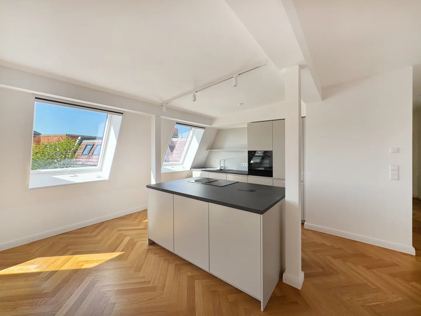 Bright, modern kitchen with white walls, herringbone wood floors, and a gray island. Skylight windows offer a view of rooftops and blue sky.