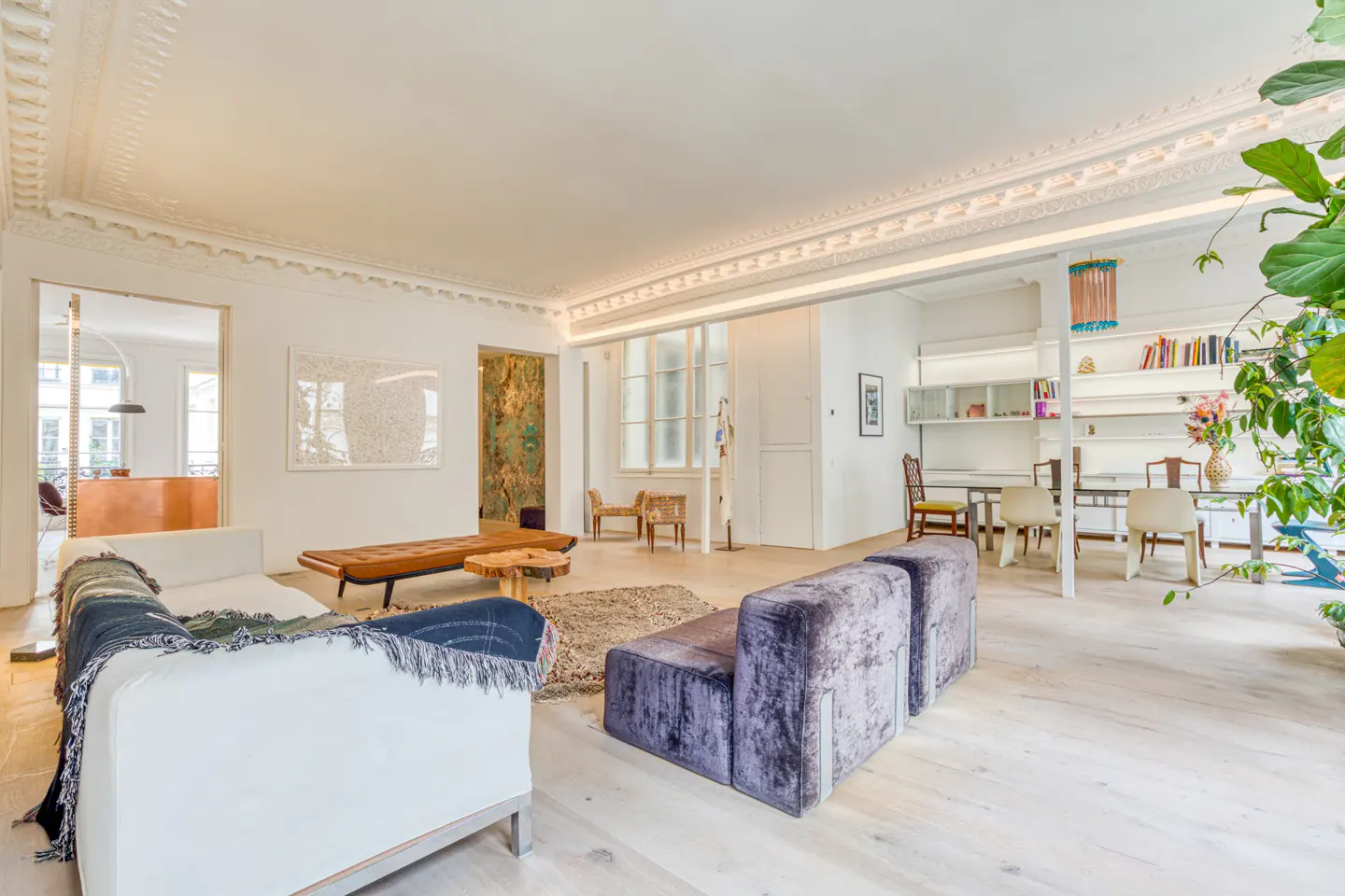 Bright, airy living room with white walls, light wood floors, and ornate ceiling trim. Features a white sofa, purple velvet couch, and dining area.
