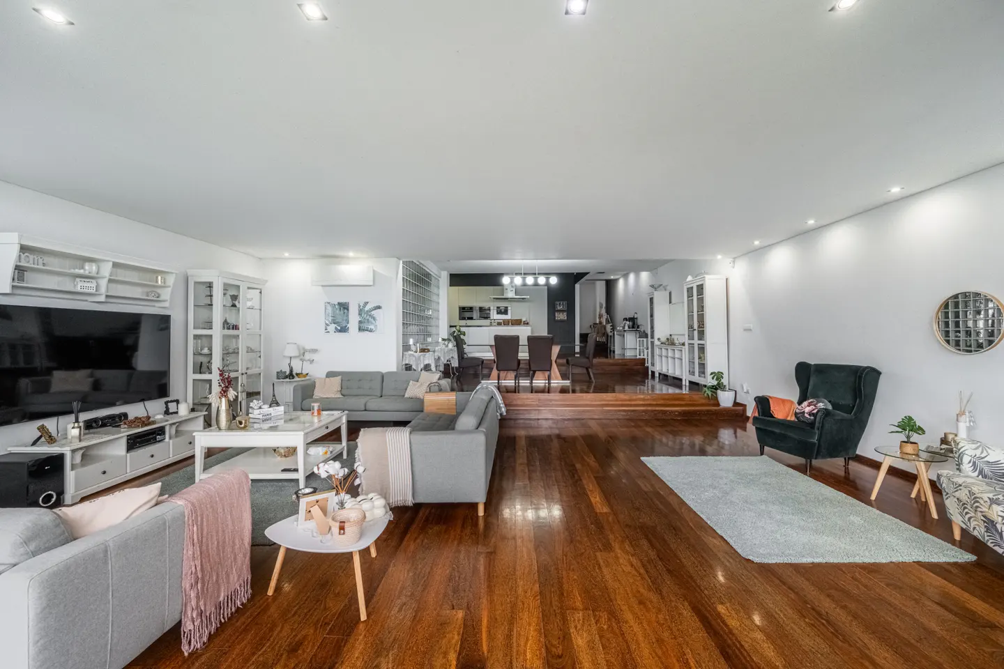 Spacious open-plan living room with dark hardwood floors, grey sofas, a black armchair, and a dining area on a raised level.