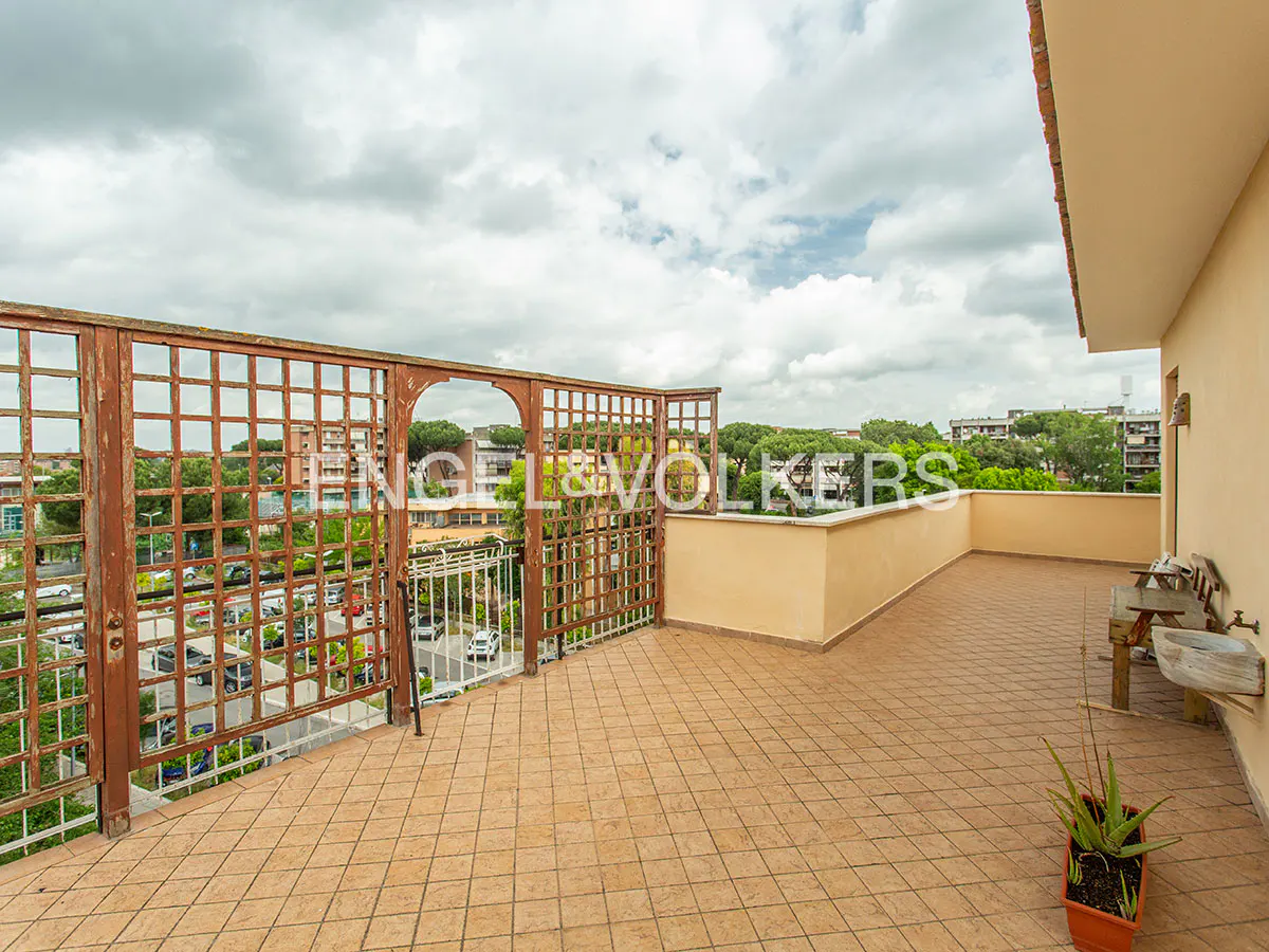 A rooftop terrace with brown tiled flooring, a wooden lattice fence, and a view of a city with trees under a cloudy sky.