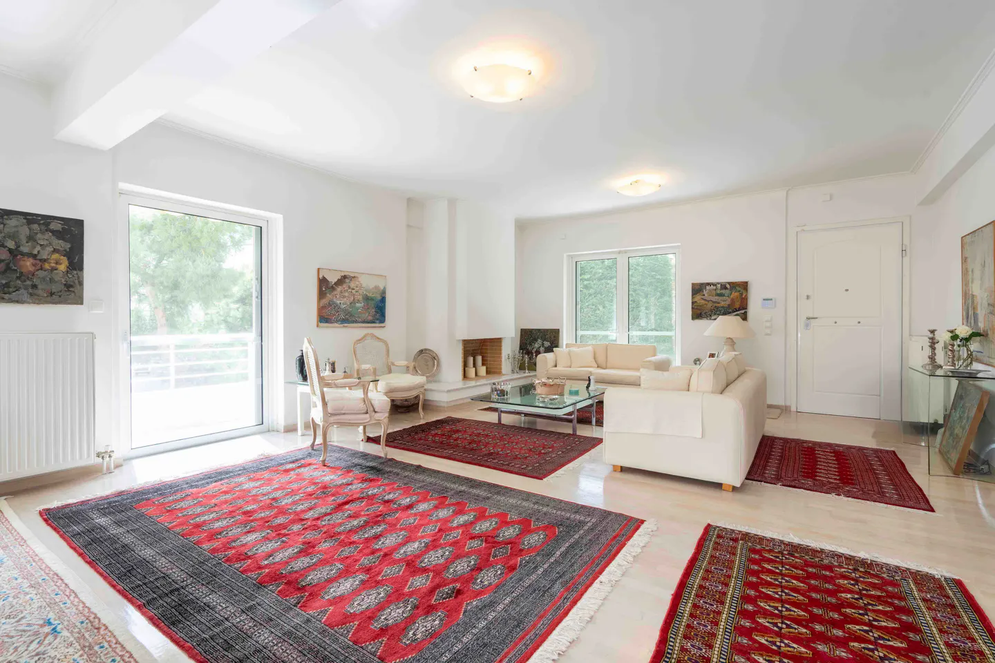Bright living room with white walls, sofas, and red patterned rugs. Art hangs on the walls, and a fireplace adds warmth.