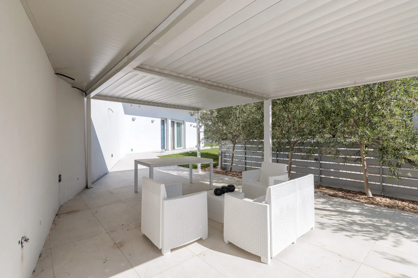 Outdoor patio with white wicker furniture under a white pergola. A white table sits in the background, with trees and a gray fence visible.