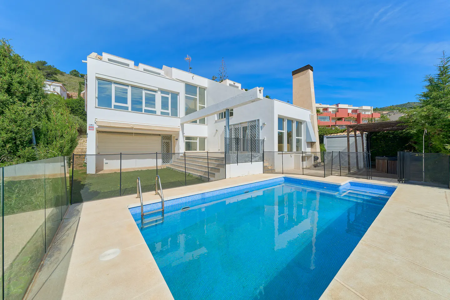 Modern white house with a blue tiled pool in the foreground under a clear blue sky.