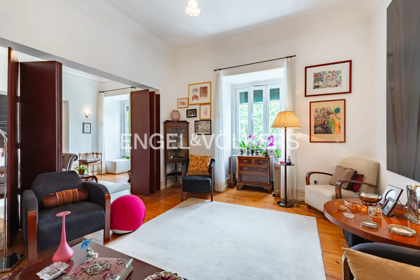 Bright living room with hardwood floors, white walls, and a large white rug. Dark armchairs, artwork, and a floor lamp add warmth.