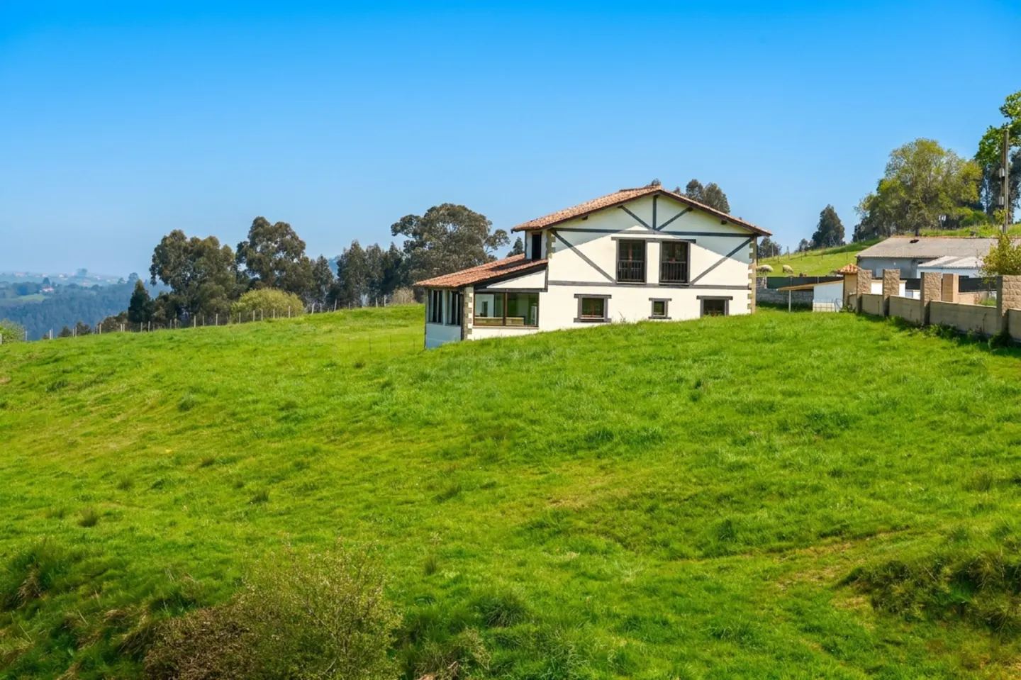 A two-story white house with a brown roof sits on a green hillside under a clear blue sky.