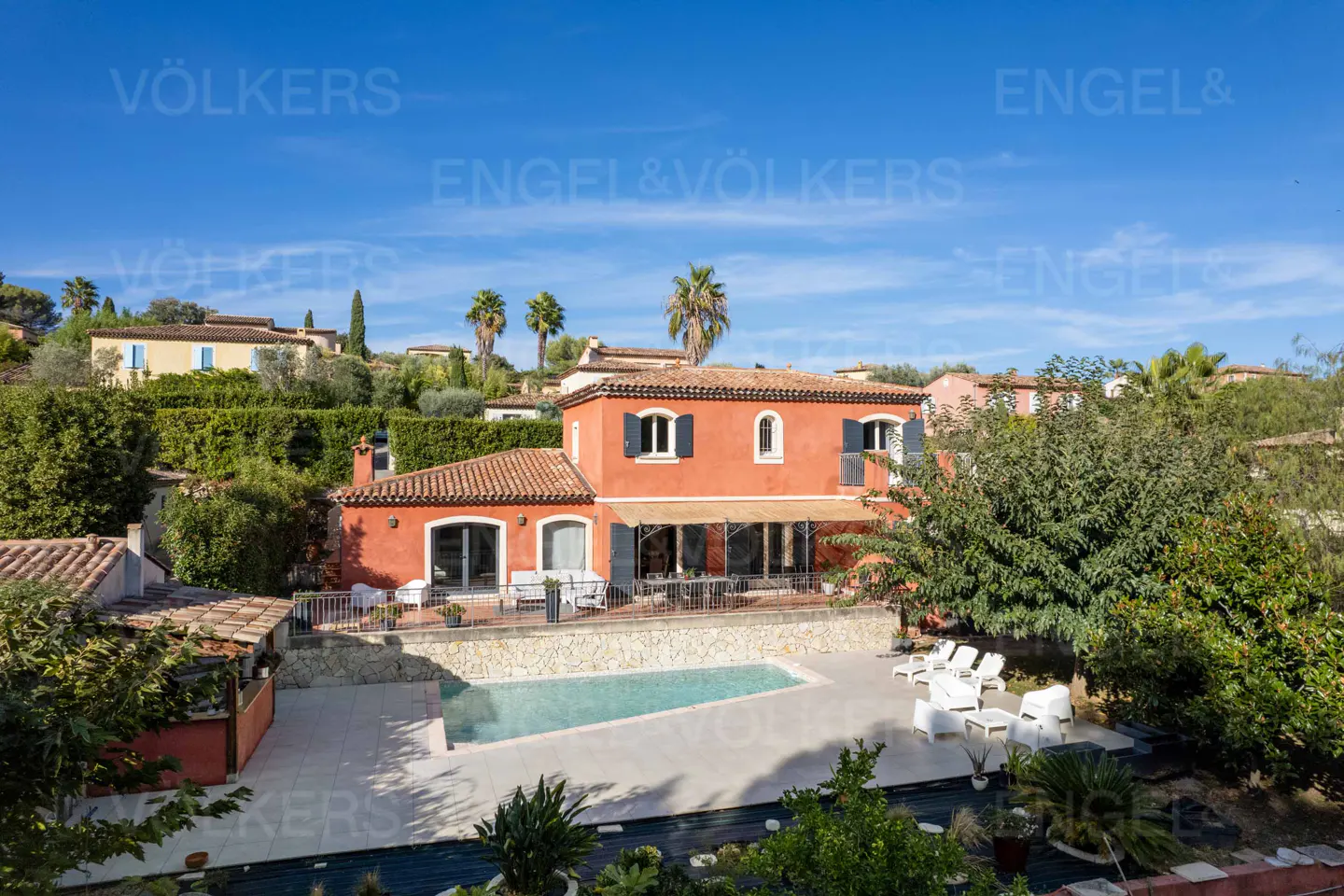 Exterior view of a two-story red house with a pool, patio furniture, and lush greenery under a blue sky.