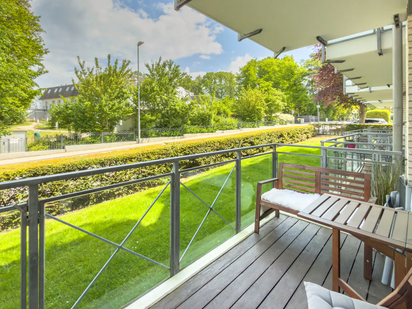 Balcony with wooden decking, a bench with a white cushion, and a small table, overlooking a green lawn and lush trees.