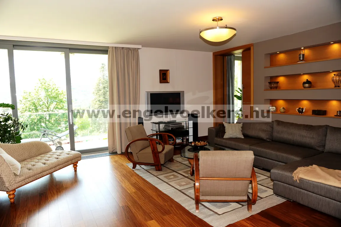 Living room with brown sofa, chairs, and wood floors. Shelves with decorative items are on the wall. Large windows overlook greenery.