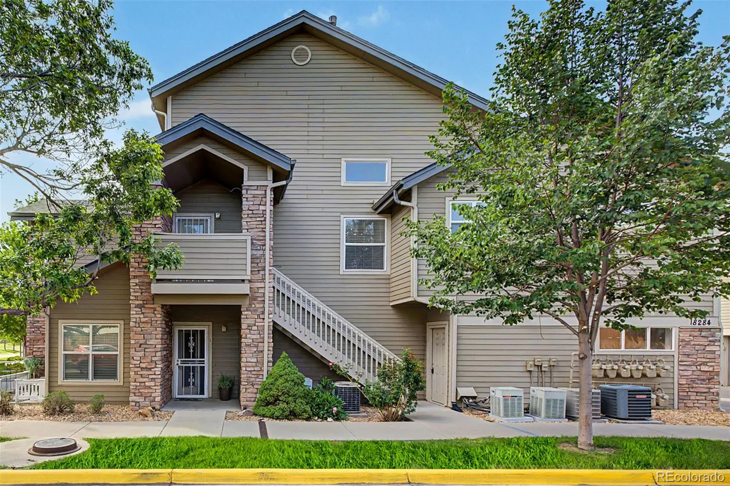 Exterior of a two-story, tan condo with stone accents, a staircase, and green trees.